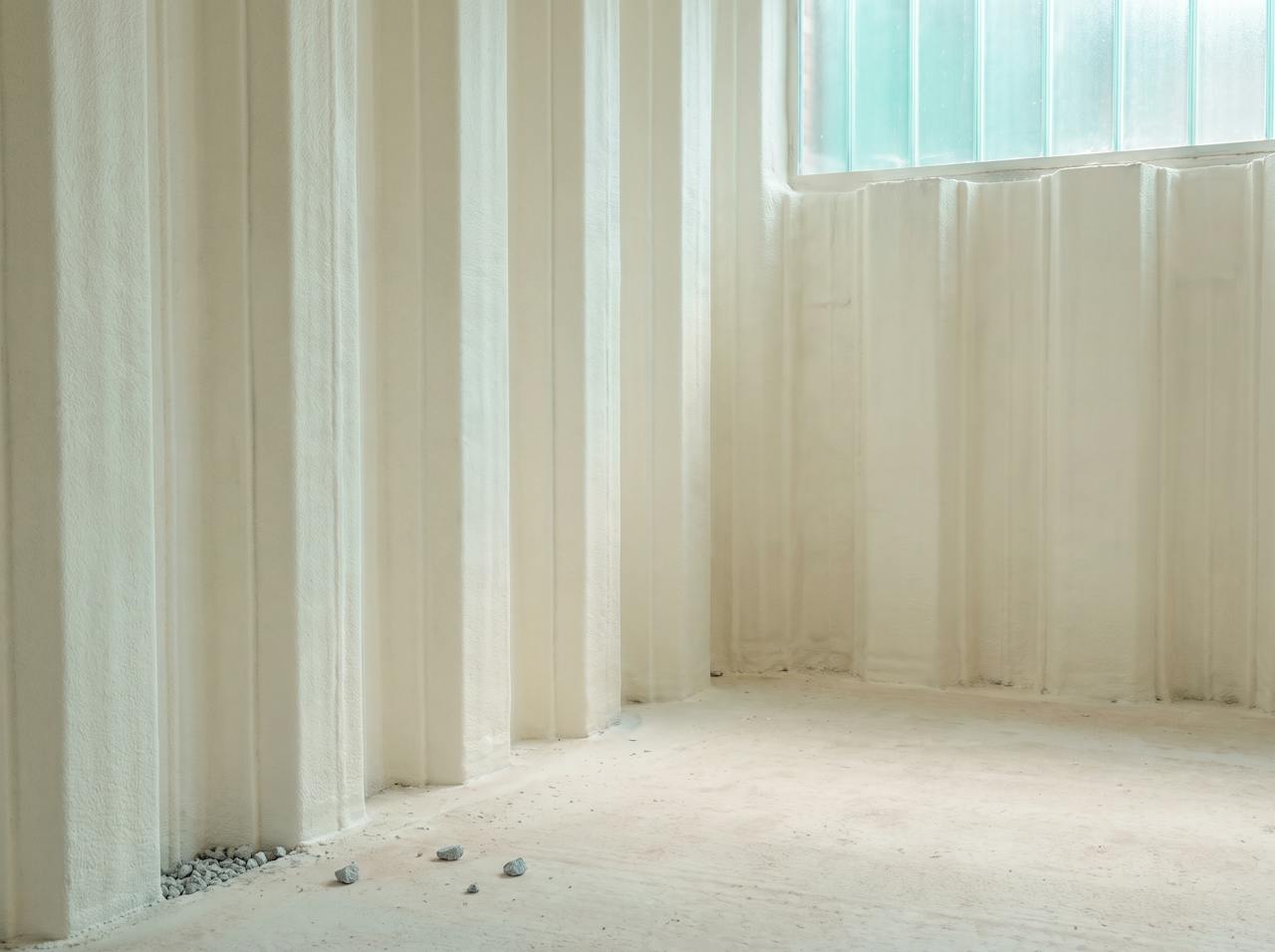 A steel sheet-piled basement wall with a continuous cream closed-cell layer filling the profile and finished to a flat face, stopped cleanly at the floor slab, soft even light.