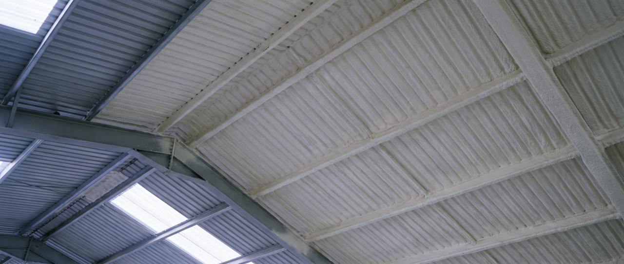 The same kind of roof from the floor: the underside of a profiled roof with a continuous pale closed-cell layer following every rib and wrapping the purlins, one bay at the left edge still bare grey sheet, the rooflights left clear.