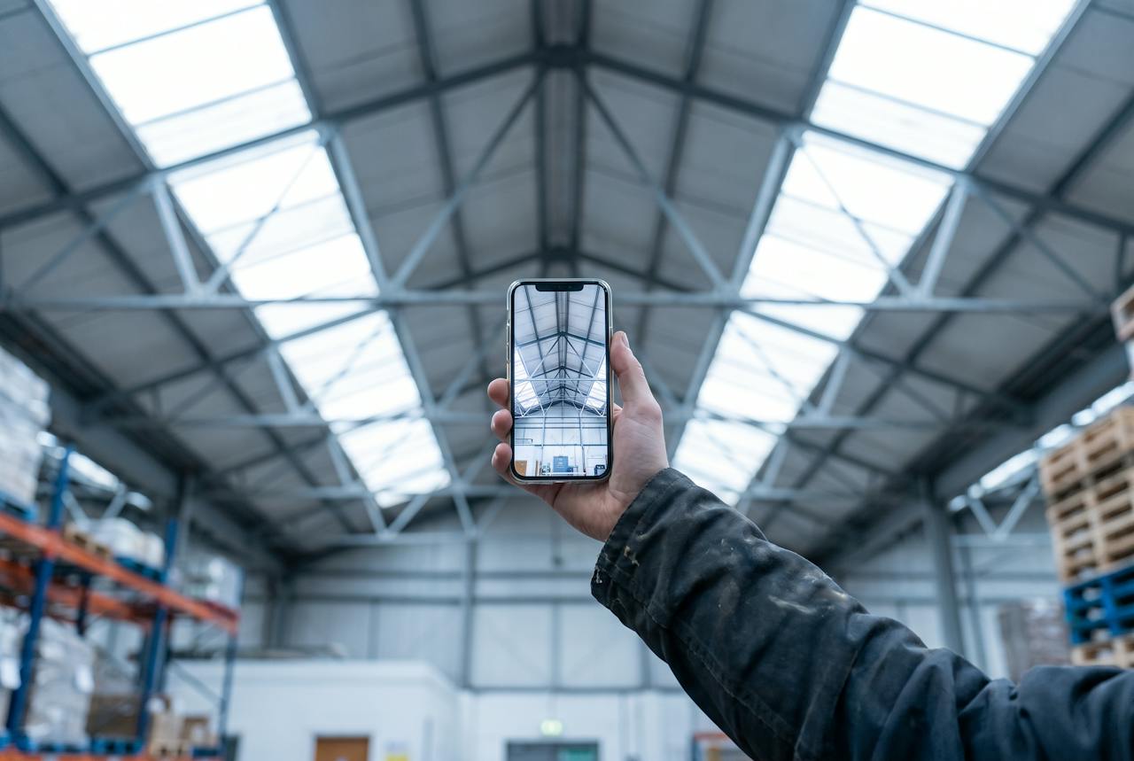 A hand holding a phone up to photograph the pale underside of a large commercial roof, purlins and rooflights in the frame.