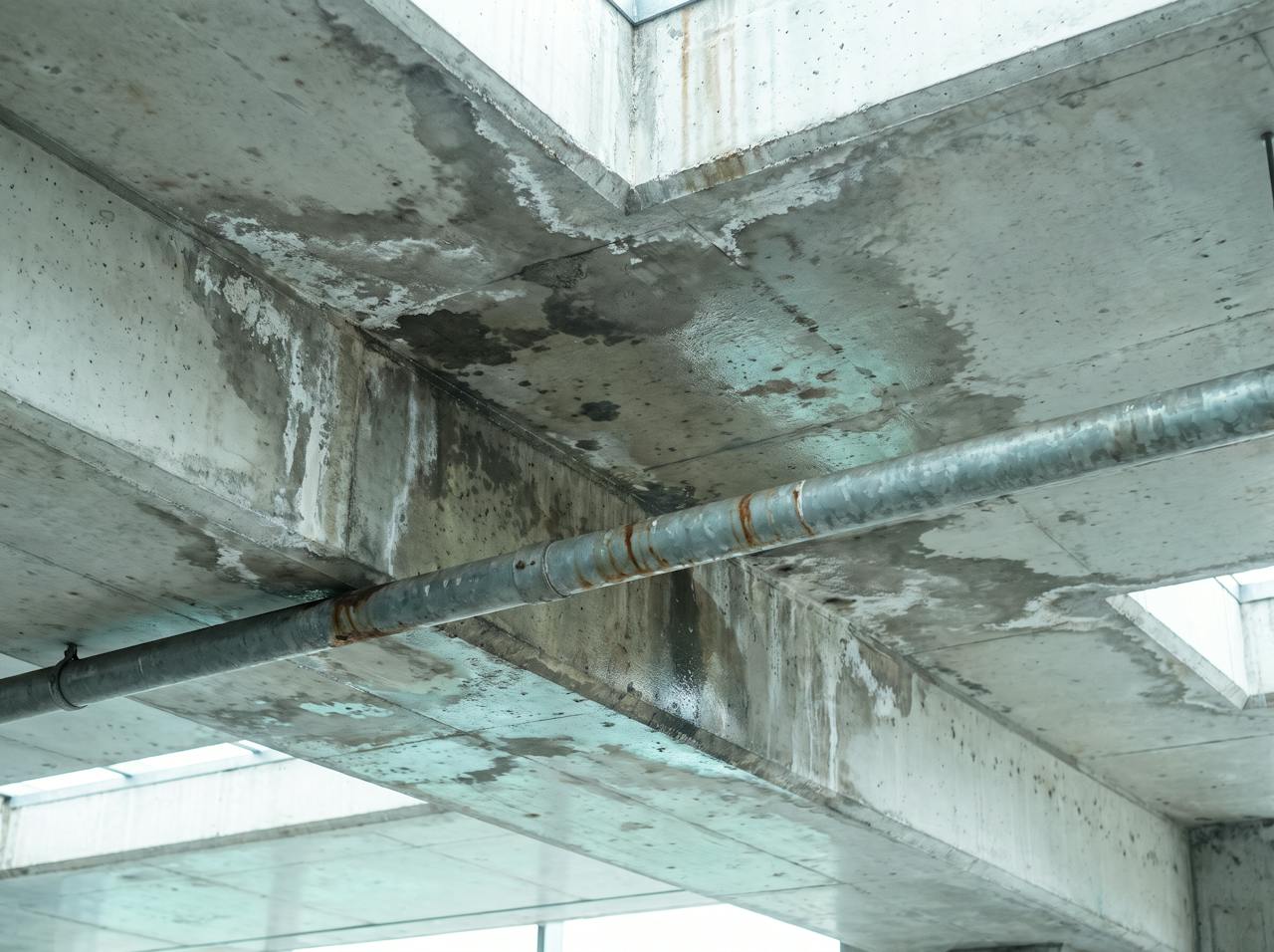 Looking up at a bare concrete car park soffit in winter: dark damp staining and condensation along a downstand beam and around a service penetration.