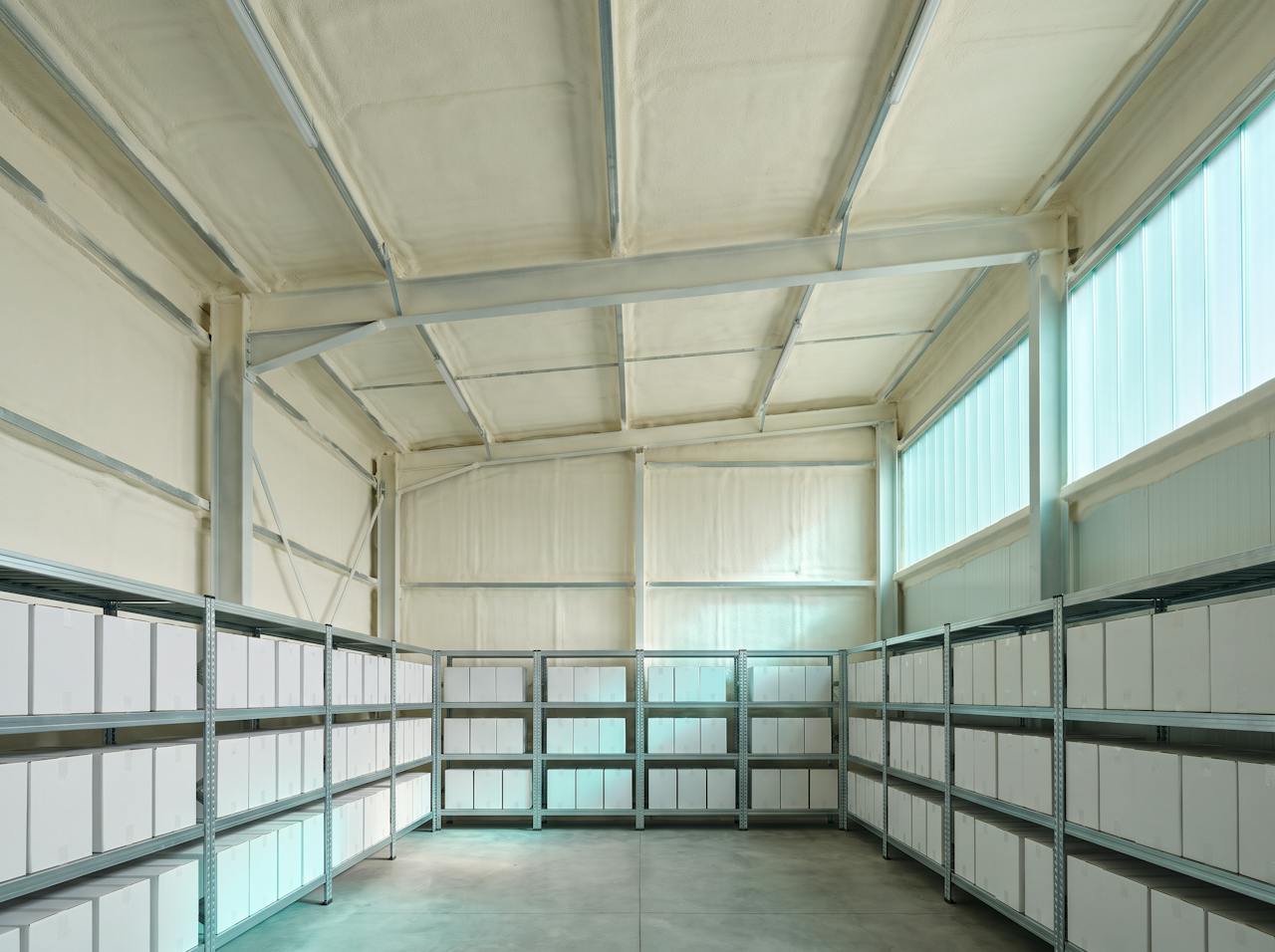 Inside a cold store roof void after the work: the underside of the roof sheet, its purlins and the top of the panel ceiling in one continuous cream closed-cell layer, chilled pipework passing through and sealed around.