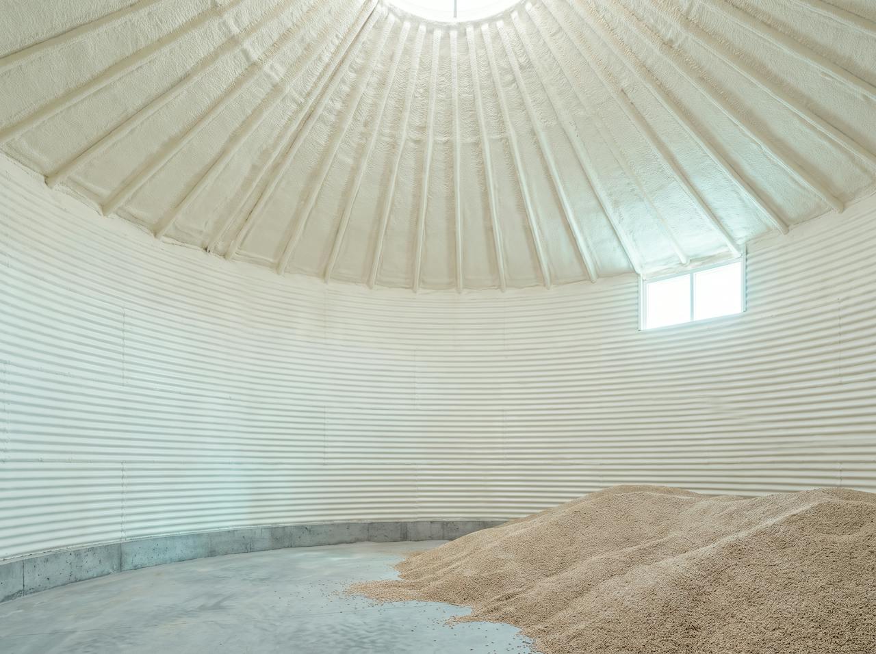 Inside a round silo after the work: the walls and conical roof sprayed in a continuous cream closed-cell layer between timber studs, three small windows letting in daylight.
