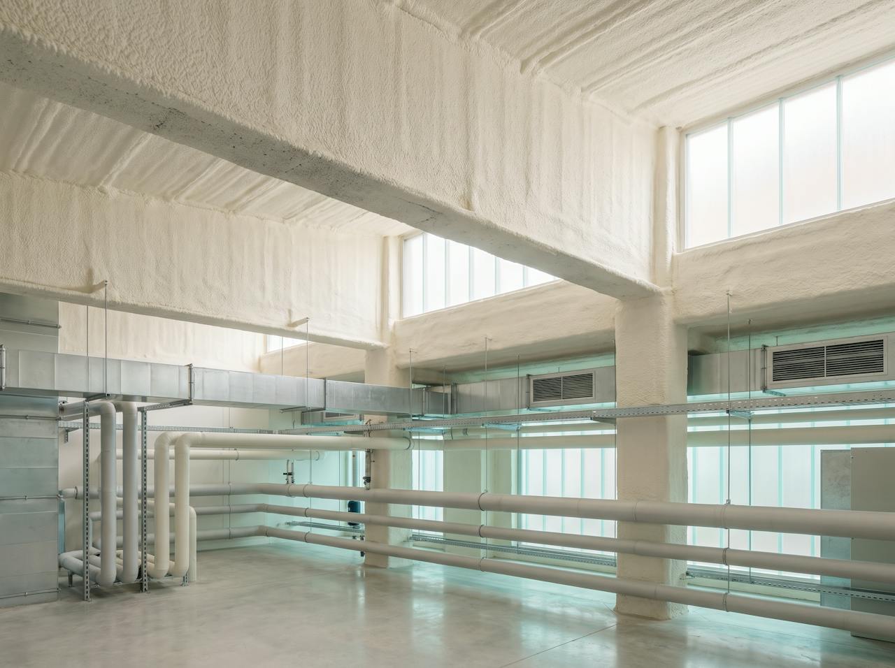 An undercroft beneath a public building after the work: the concrete soffit and its downstand beams in a continuous cream closed-cell layer, cable trays and pipework passing beneath and sealed around, cars parked below.