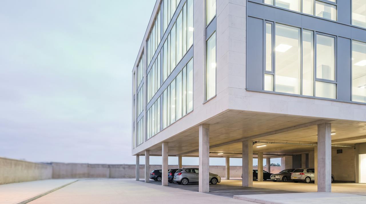 A modern UK office building at dusk with an open undercroft car park beneath it, the bare concrete soffit and its downstand beams visible above the parked cars.