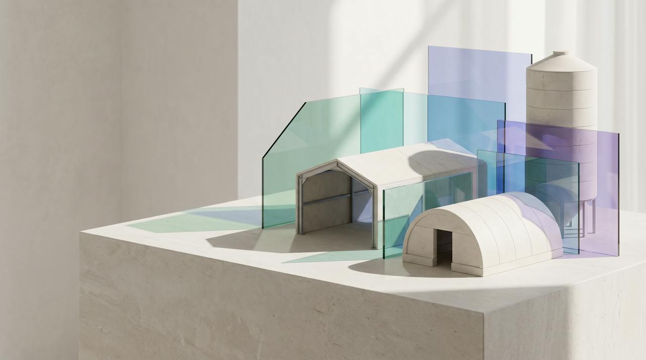 An architectural model of several building forms in pale stone, a portal frame, an arched hut and a silo, with translucent glass planes casting sea-glass, aqua and violet light across them.