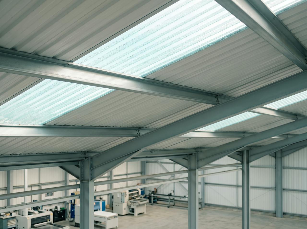 Looking up inside an industrial unit on a cold morning: the underside of a profiled steel roof misted with condensation between the purlins, a run of strip lights, and the tops of machines and finished product below.