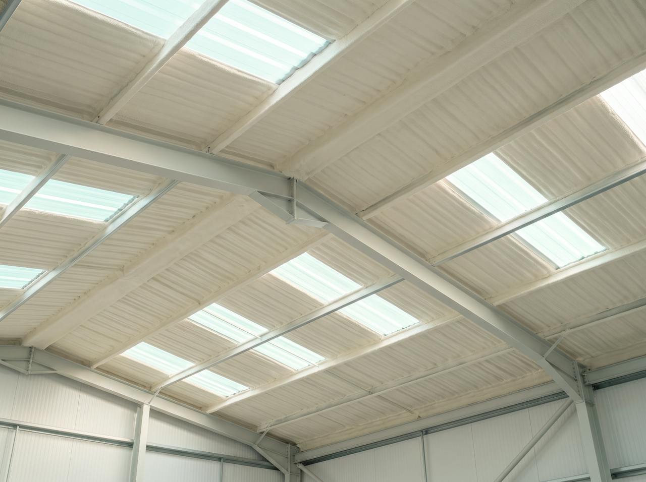 Looking up inside a factory unit after the work: the underside of the profiled steel roof and its purlins in a continuous cream closed-cell layer, strip lights left clear, machinery back in place beneath.