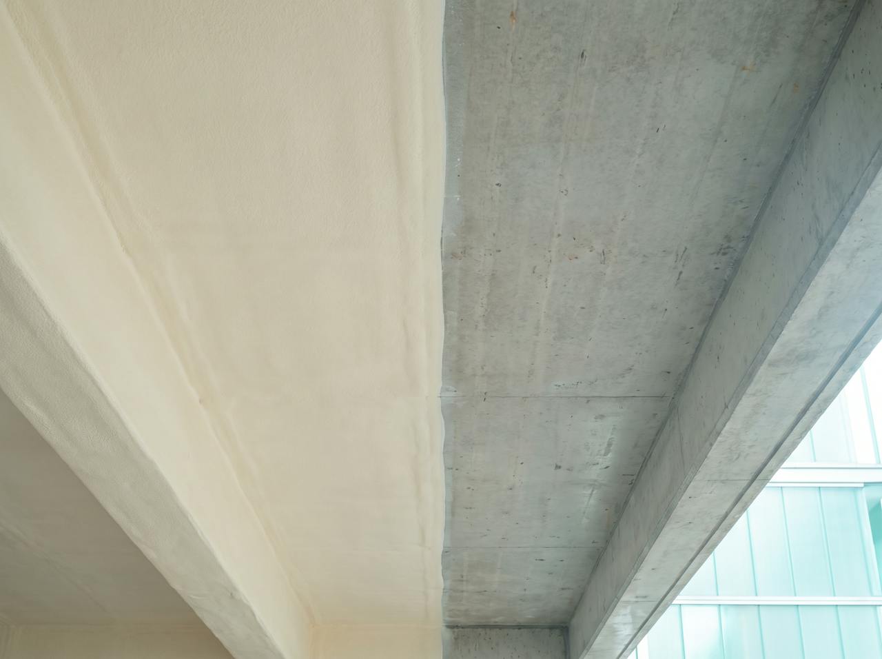 The concrete soffit of an undercroft car park, one half still bare grey concrete, the other half covered in a continuous cream closed-cell foam layer wrapping a downstand beam.