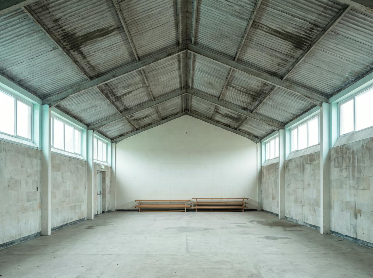 Inside a school sports hall on a cold morning: condensation beaded on the underside of the profiled steel roof between the trusses, a drip mark spreading on the court floor below and equipment stacked against the wall.