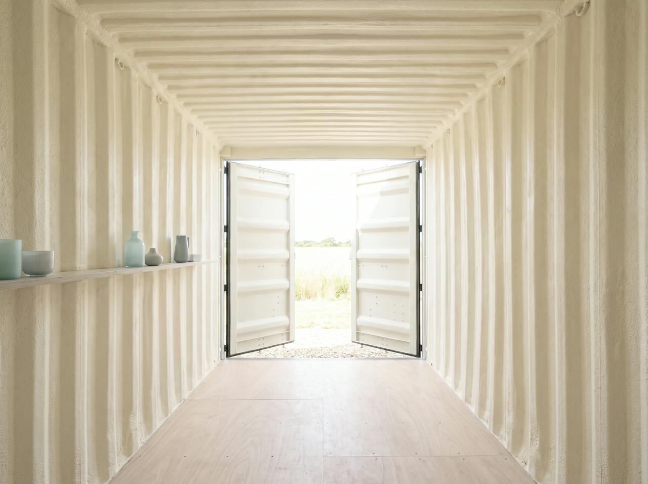 Inside a shipping container after the work: walls and roof sprayed in a continuous cream closed-cell layer following the corrugation, timber battens fixed ready for the lining, daylight through the open doors.
