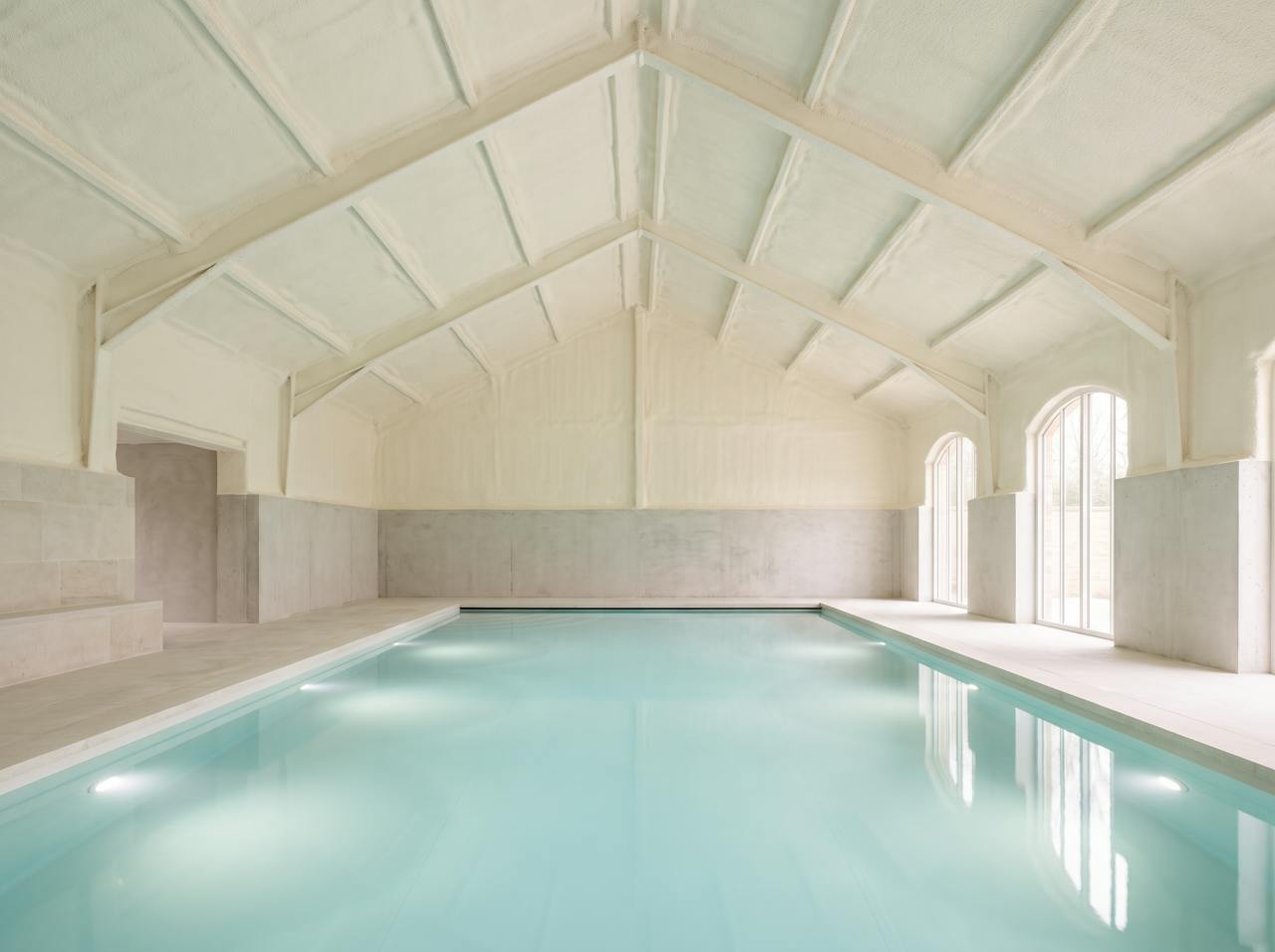 Inside a pool hall roof after the work: the underside of the deck and its beams in a continuous cream closed-cell layer, the lighting rig and ventilation grilles left clear above the water.