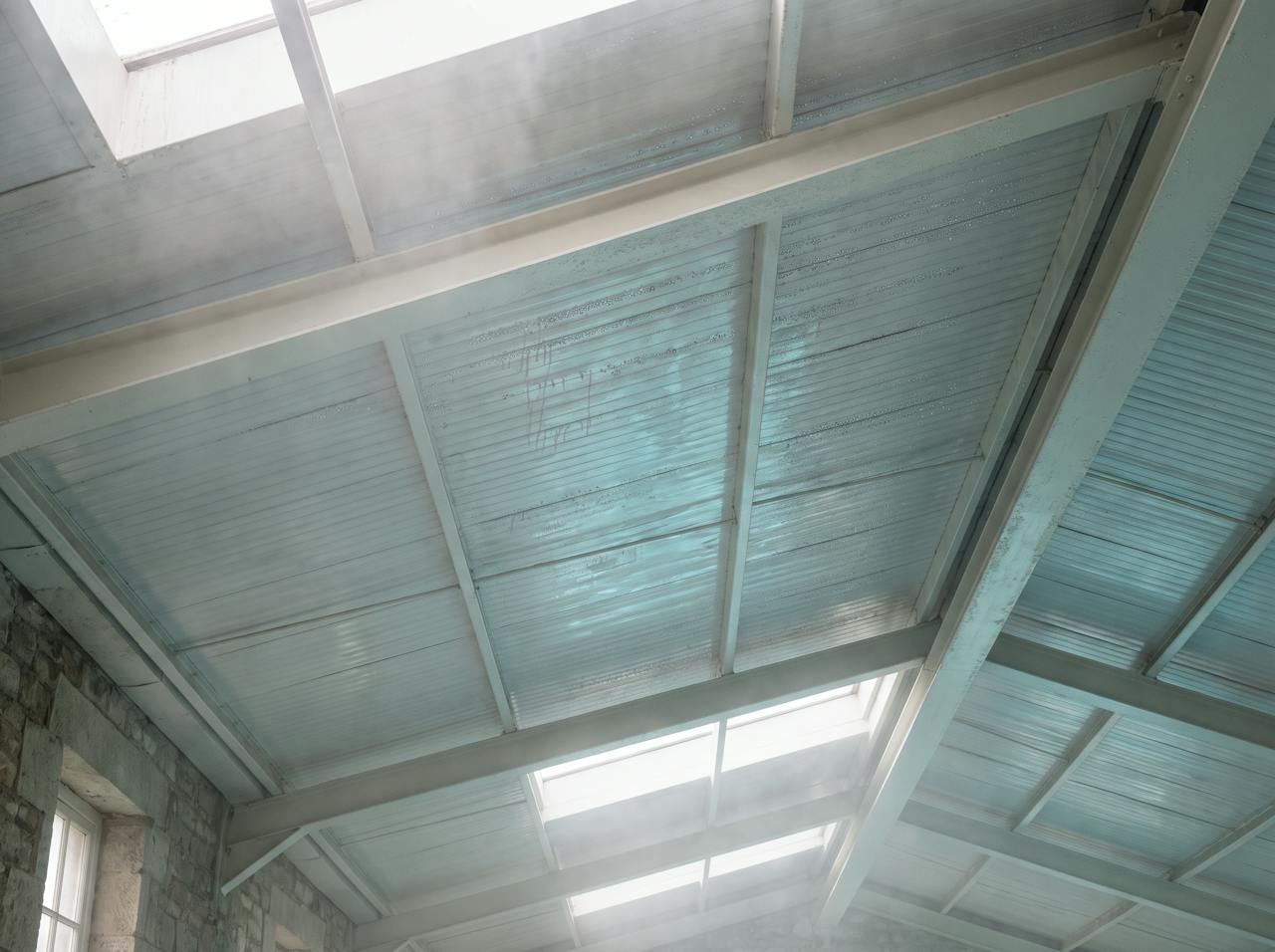 Looking up inside a pool hall: condensation beaded along steel purlins and the underside of a roof deck, dark staining spreading from the fixings, and a drip line on the poolside tiles below.