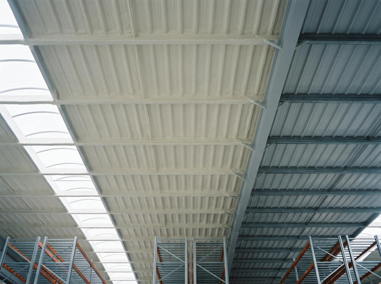 Looking up inside a distribution warehouse: the underside of a profiled steel roof after a closed-cell layer has been applied from below, following the ribs and wrapping the purlin flanges, meeting the next bay of bare grey steel at a straight line along a rafter.