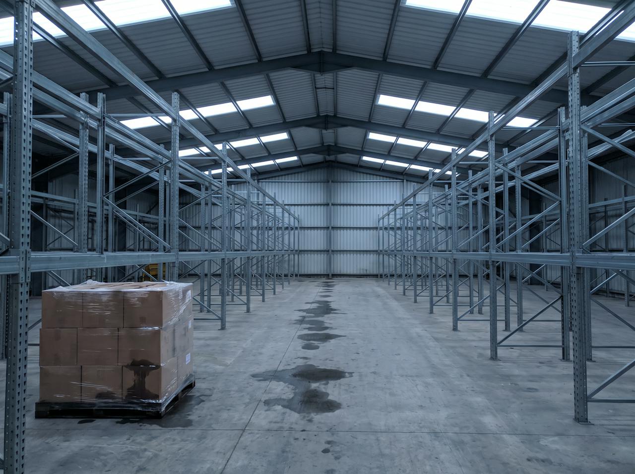 A wide aisle between tall pallet racking in a distribution warehouse, with a line of dark wet patches along the concrete floor beneath one valley of the roof and a shrink-wrapped pallet of cardboard boxes with darkened wet corners.