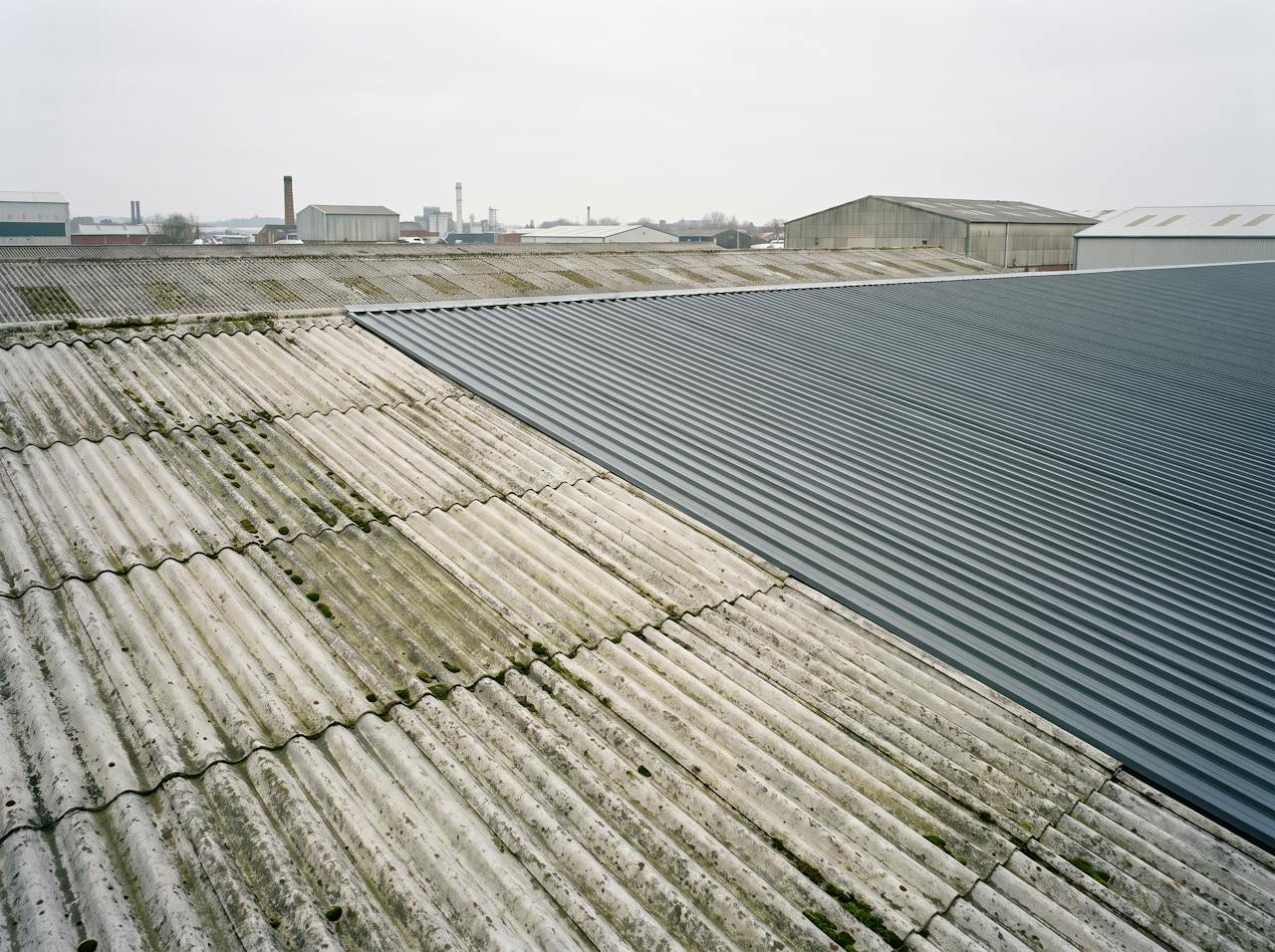 Across the roof of a large single-storey industrial building on an overcast day: an old profiled fibre-cement roof, weathered with moss in the troughs, meeting a bay overclad in new dark grey profiled steel at a straight line.