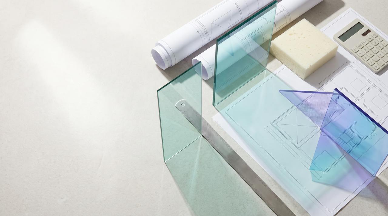 Seen from above: roof plan drawings on a pale desk with a steel rule, a cream closed-cell foam sample block and a calculator, lit by coloured light through glass planes.