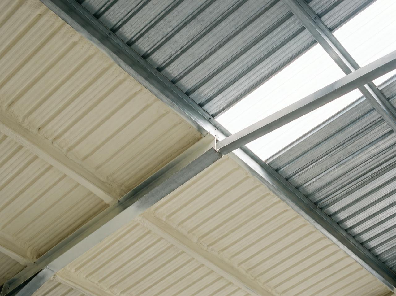 The underside of a profiled roof seen from the floor: the treated bays in a continuous cream layer, the untreated bays still bare grey steel, and a crisp boundary between them beside a rooflight.