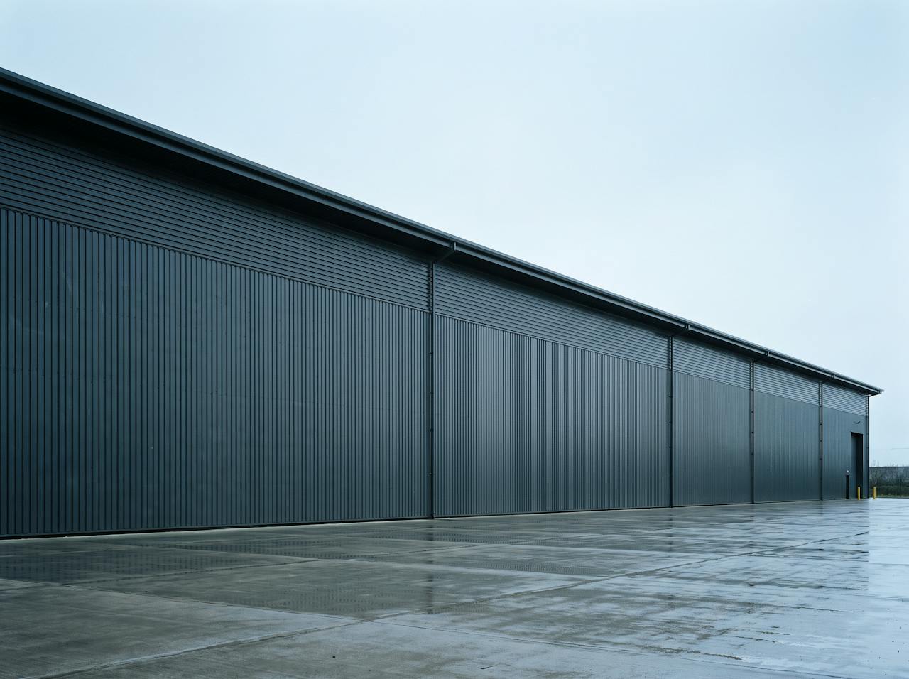 A long, low distribution warehouse in dark grey profiled cladding under an overcast sky, with a wet concrete apron in front.