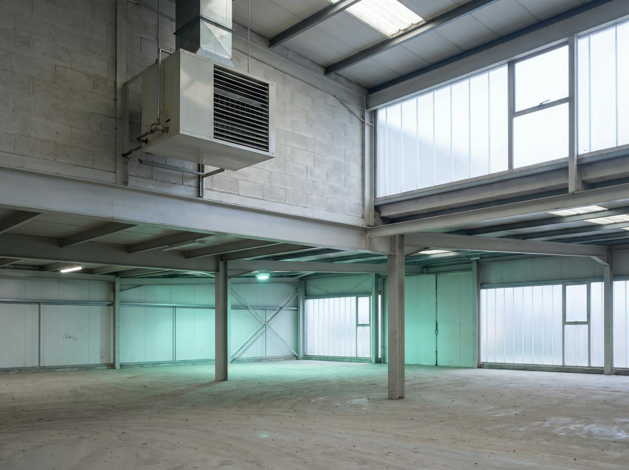 Inside a cold warehouse on a winter morning: a suspended warm-air heater running beneath a bare profiled steel roof, breath visible, racking in the background.
