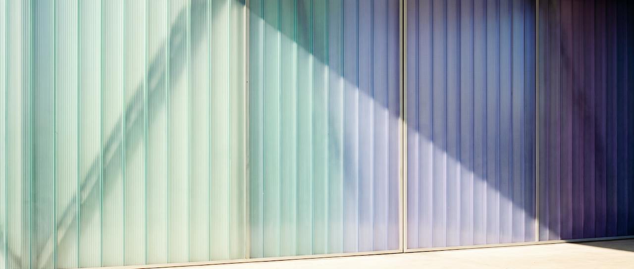 Daylight passing through a wall of fluted translucent panels, graded from sea-glass green through aqua and periwinkle to violet, with the shadows of the structure cast across the ribs.