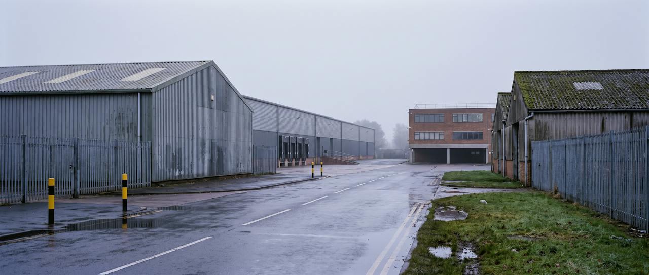 A mixed British industrial estate on a cold, misty morning seen along a wet estate road: a steel-framed unit in weathered grey profiled cladding with rooflight strips in the foreground, a long distribution warehouse with a row of dock doors behind it, a brick two-storey office block at the end of the road and an older unit with a mossy corrugated fibre-cement roof on the right, yellow-and-black bollards and palisade fencing.