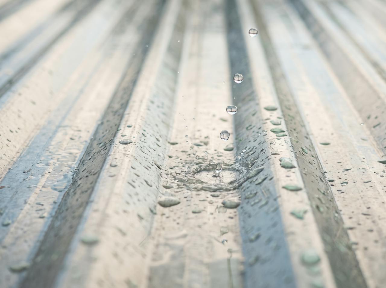 Looking up at a bare profiled steel roof from inside a workshop during heavy rain, water streaming down the rooflight, the thin sheet spanning between purlins.