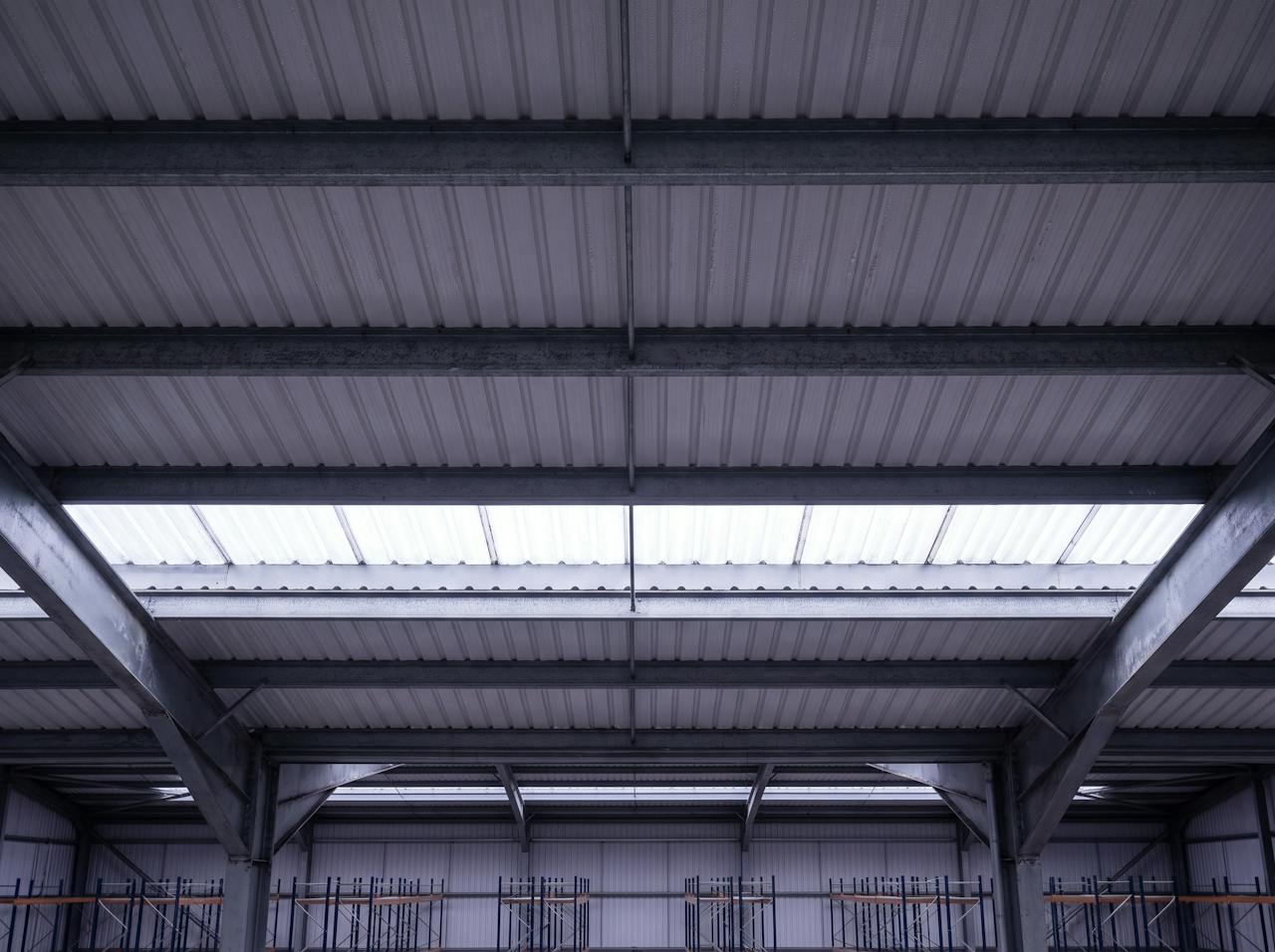 Looking up inside a distribution warehouse on a cold morning: the bare underside of a grey profiled steel roof sheet between galvanised purlins, fine condensation across the sheet, a translucent rooflight strip, steel rafters and the tops of pallet racking below.