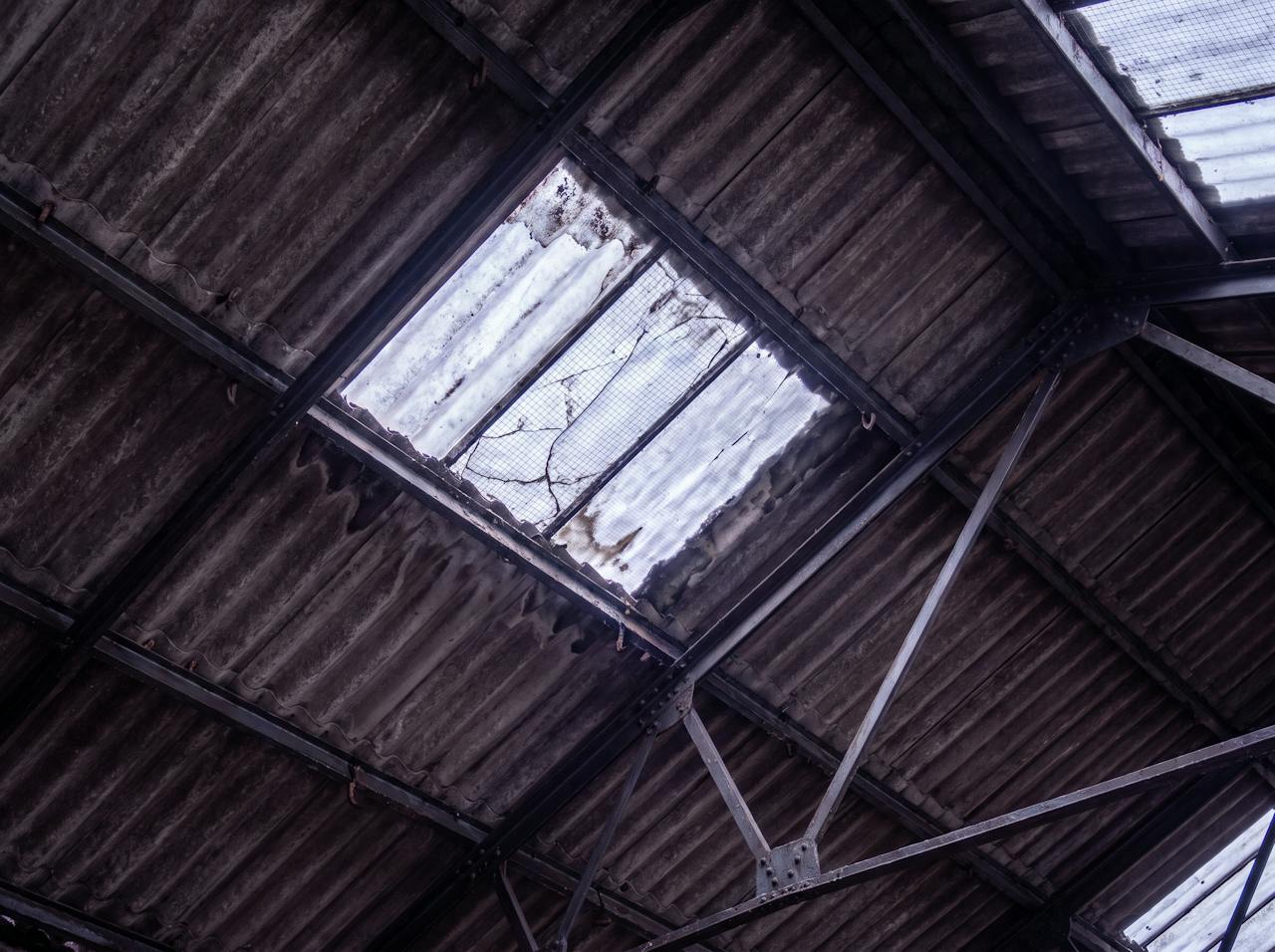 Looking up inside an old factory unit on a cold grey morning: the underside of a pitched roof of dark, weathered wide-profile asbestos-cement sheets with hook bolts, riveted steel lattice trusses, and a cracked wire-reinforced rooflight.