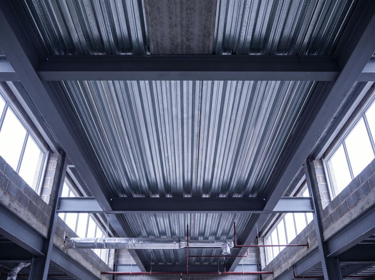 Looking up inside a new-build warehouse before fit-out: the bare underside of a composite steel and concrete roof deck, galvanised trapezoidal decking ribs between steel beams, ductwork and sprinkler pipework hung beneath it, daylight from high-level windows.
