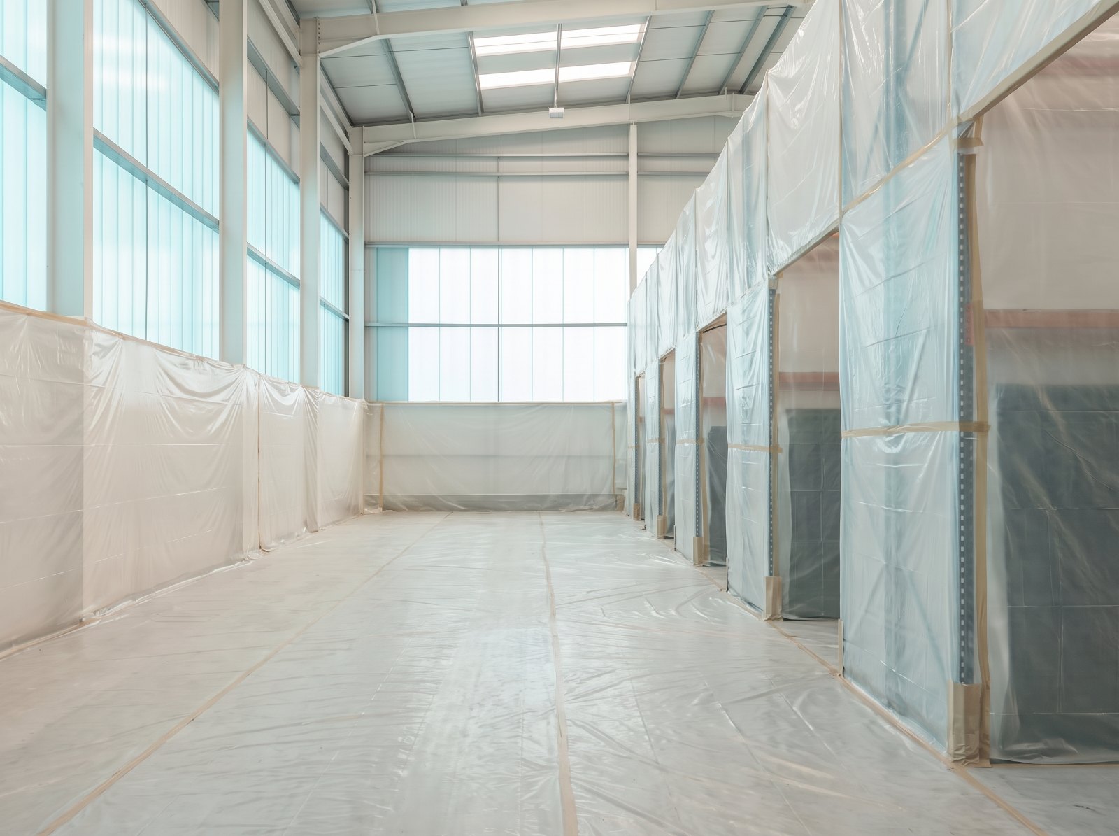 A live warehouse interior prepared for spraying: polythene sheeting hung between the bays, racking and stock covered, rooflights and services masked.