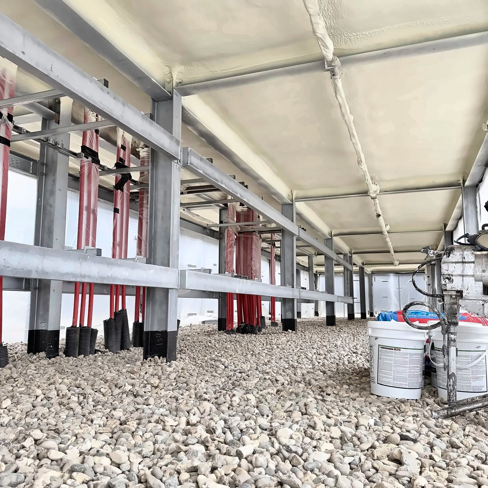 A concrete deck soffit over an undercroft void fully sprayed in a continuous cream closed-cell layer, galvanised steel frame and red cable ducts below it, NexSeal drums and the spray gun on the gravel floor.