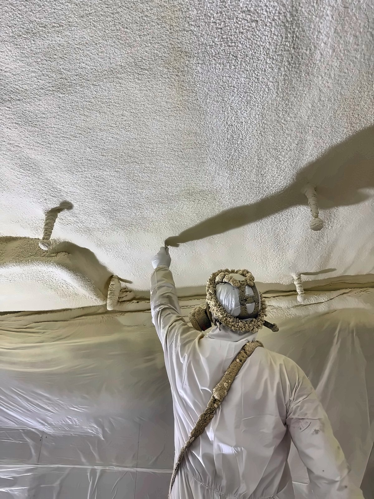 A HOLVEM installer in full protective clothing spraying closed-cell foam onto an overhead soffit, with polythene masking hung below the work.