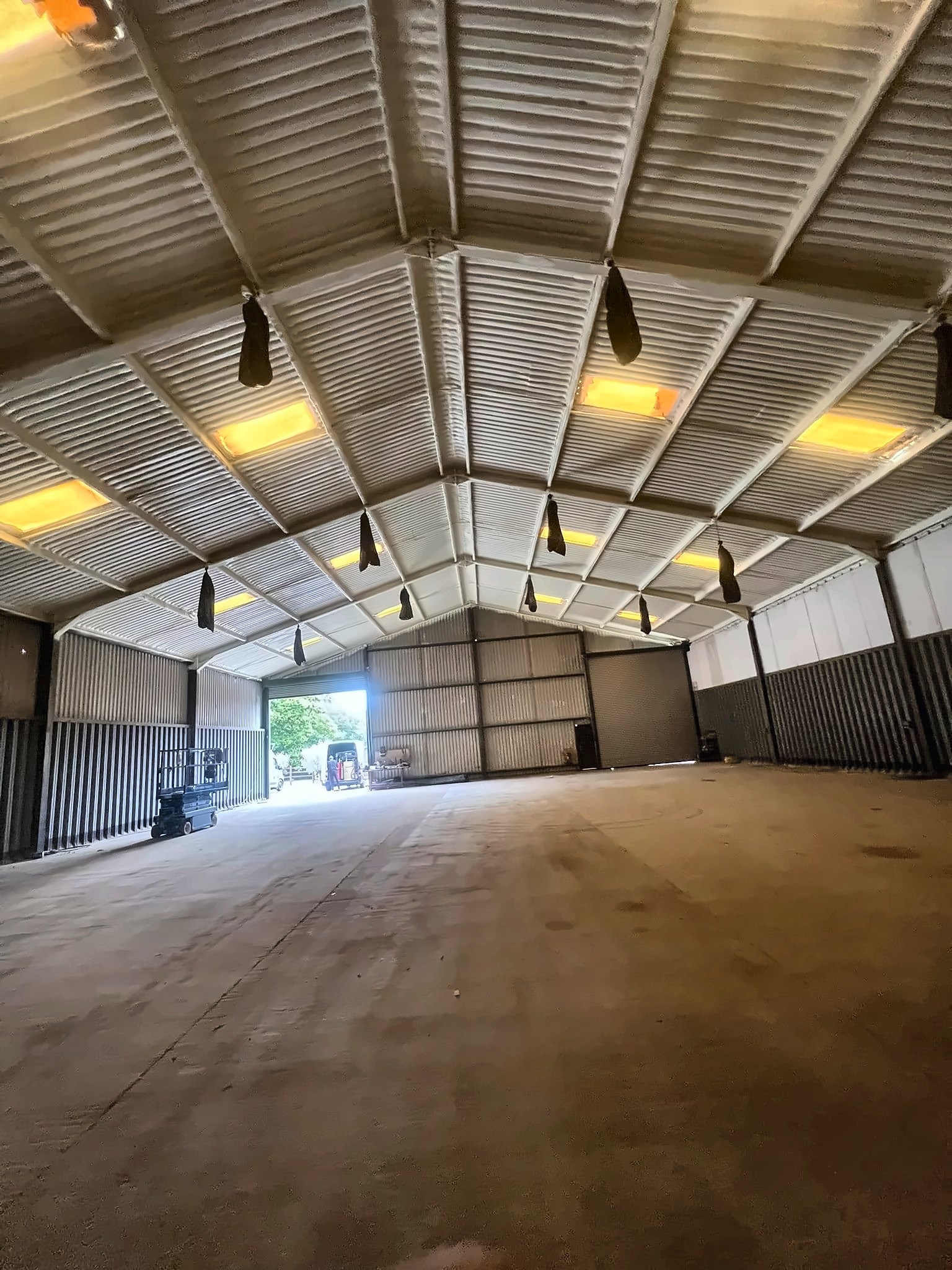 A steel portal-frame building with its roof fully sprayed from beneath, warm lights and an open door at the far end.