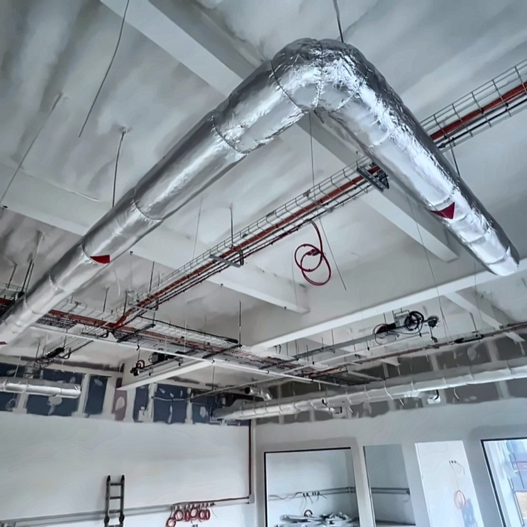 A concrete roof deck soffit sprayed in a continuous cream closed-cell layer above a new-build commercial interior, with foil-lagged ductwork and cable trays running beneath it.