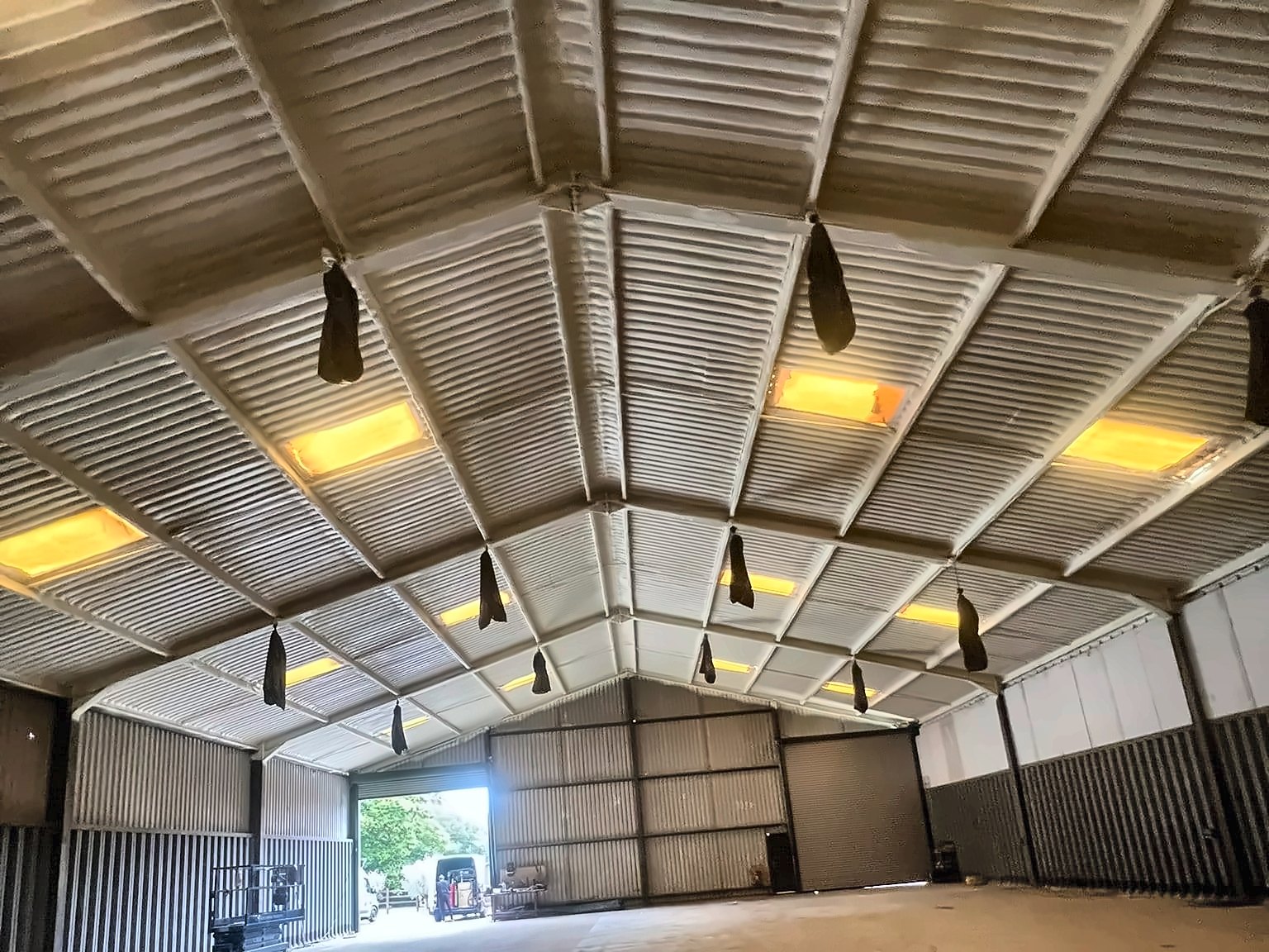 A finished job: the whole pitched steel roof of a large portal-frame building sprayed from beneath in a continuous cream closed-cell layer, purlins and rafters wrapped, the doors open to daylight.