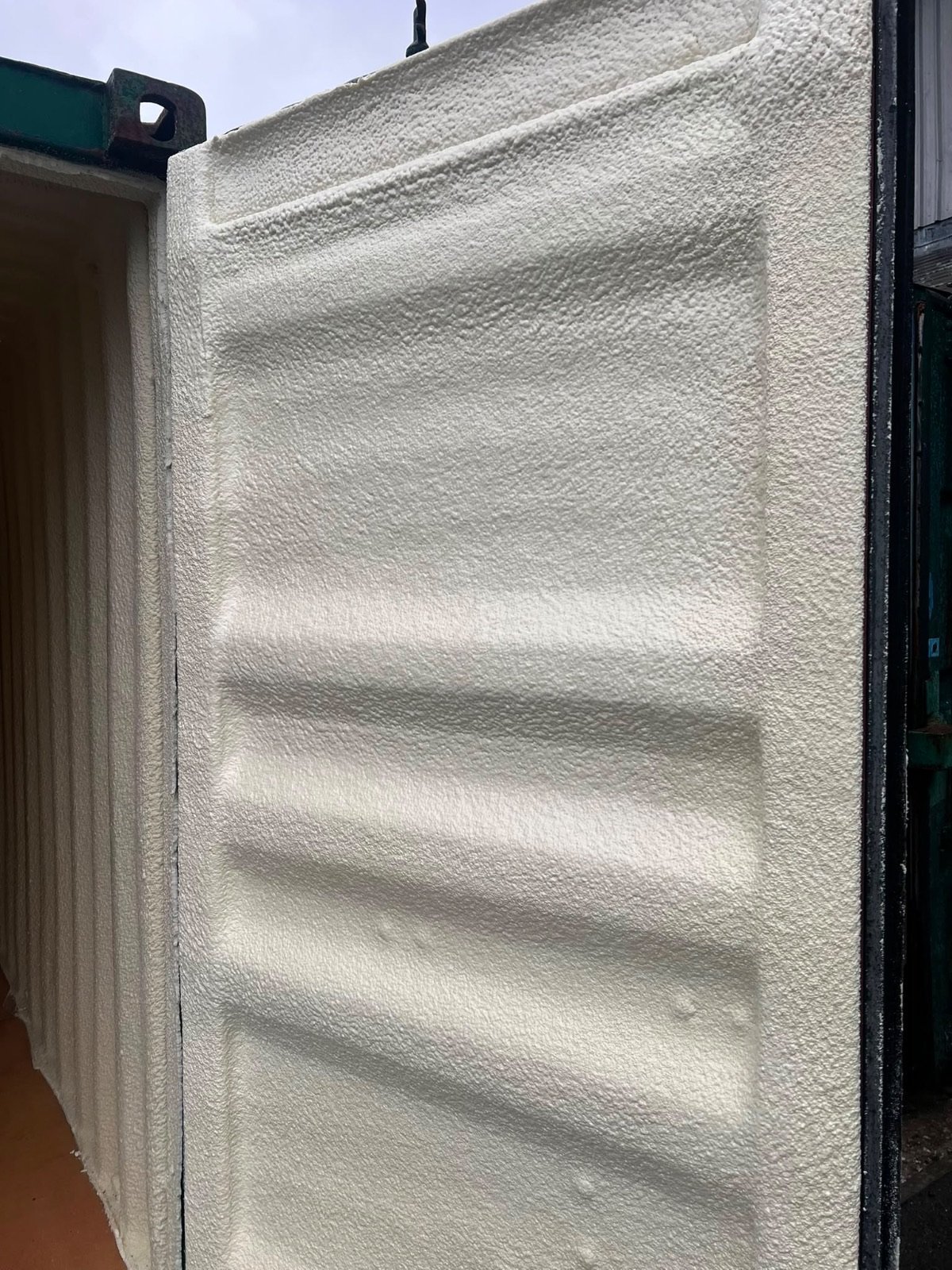 The inside of a container door leaf after the work: the corrugated steel of the door in a continuous cream closed-cell layer, the locking bars left clear.