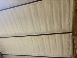 Close to the roof after the work: the corrugated sheet between two timber purlins covered in a continuous cream closed-cell layer that follows every rib, the purlins left exposed and a lamp fitting below.