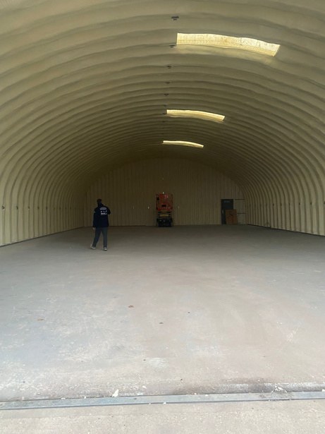 Inside the Nissen hut with the foam on and the coating still to come: the whole arch in a continuous cream closed-cell layer, three rooflight strips glowing along the crown, a person walking across the empty floor.