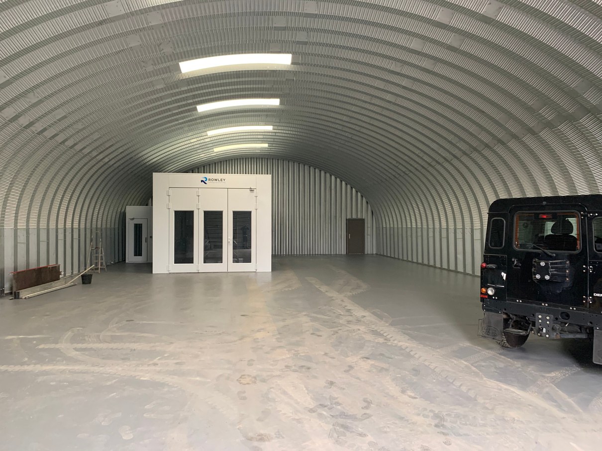 The Nissen hut before the work: bare galvanised corrugated steel from floor to crown, rooflight strips along the top, a white spray booth at the far end and a black four-wheel-drive parked inside.