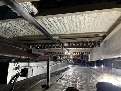 Looking up in the roof structure above the stage: the deck between deep steel beams covered in a continuous white open-cell layer, a row of lighting-bar pulleys along one beam and polythene sheeting over the grid below.