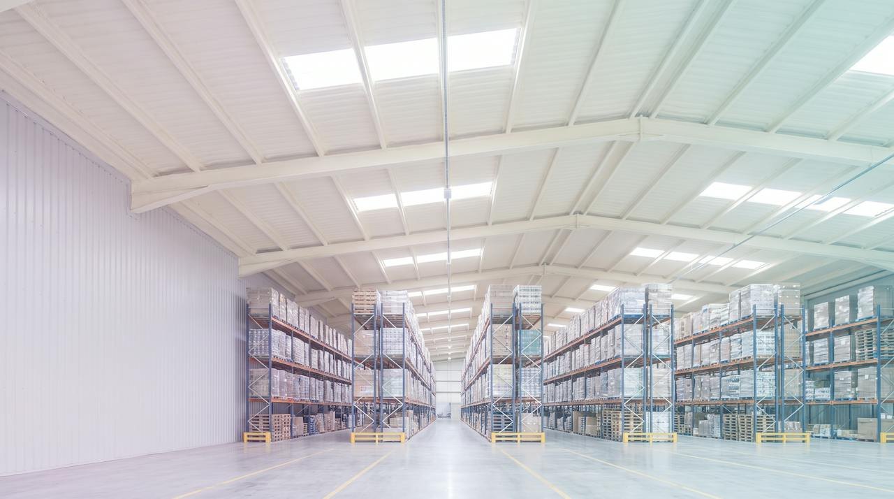 Inside a large distribution warehouse in pale morning light: tall pallet racking down a wide aisle under a high profiled steel roof, its underside and purlins sprayed in a continuous ivory closed-cell layer, rooflights letting in soft daylight.