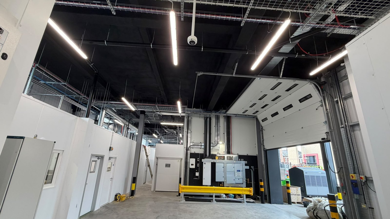 Inside a food-sector unit in London after the work: the underside of the mezzanine deck in a continuous black finish over the insulation, cable trays and lighting below, white rooms along the left and a roller door open to the yard.