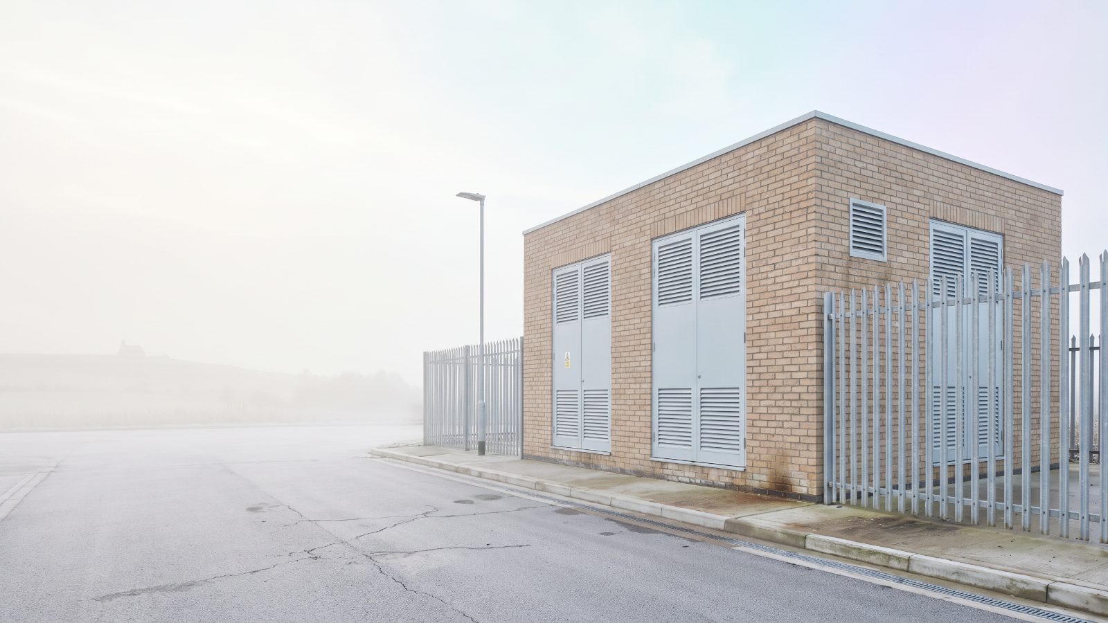 A buff brick electricity substation with louvred doors behind a palisade fence, in the mist.