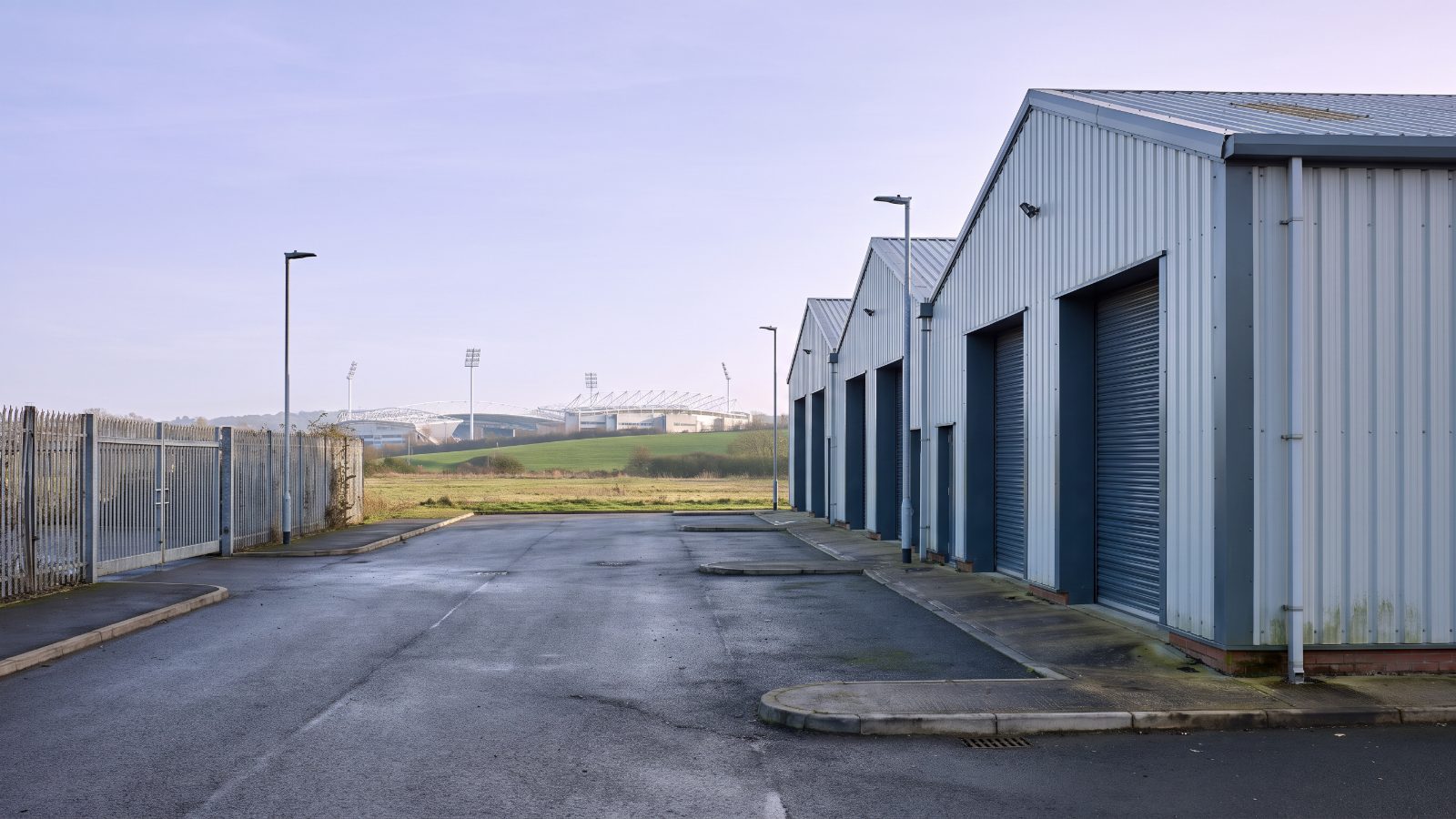 A small gated multi-let estate of pale steel portal-frame units with grey roller shutters, a security fence and gate at the left edge, a low green hillside. The tall white masts of a large city stadium stand on the skyline.