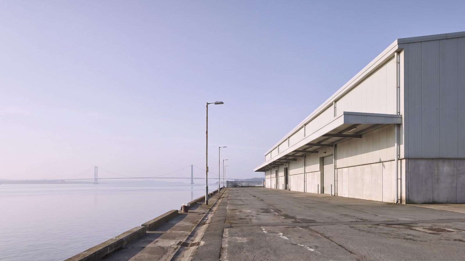A long low general goods warehouse on a port estate, pale cladding, a quay edge in front, the flat wide estuary beyond with the towers of a long suspension bridge.
