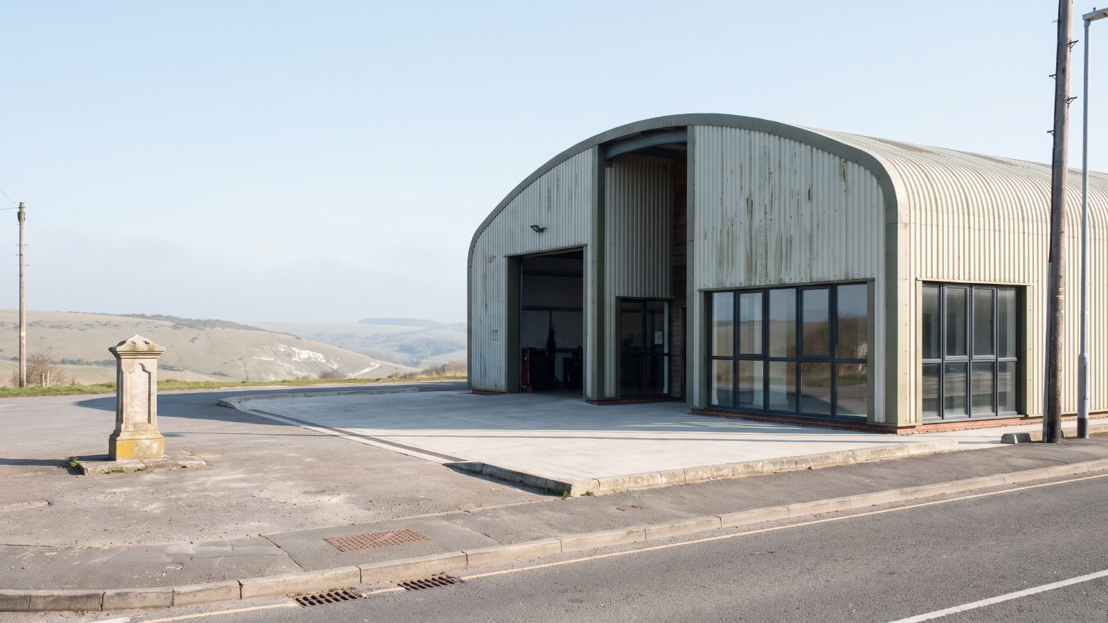 A curved-roof MOT garage workshop in pale corrugated steel with an open service bay, a quiet forecourt, chalk downland.
