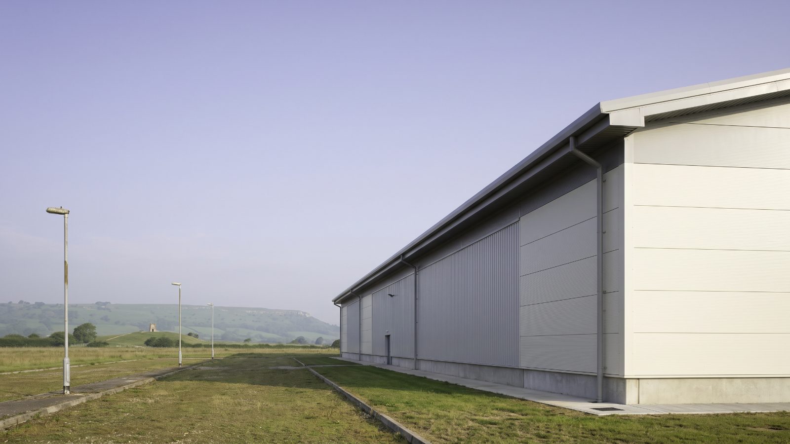 A new big-box warehouse with pale cladding and a long roofline, the Cotswold escarpment rising green.