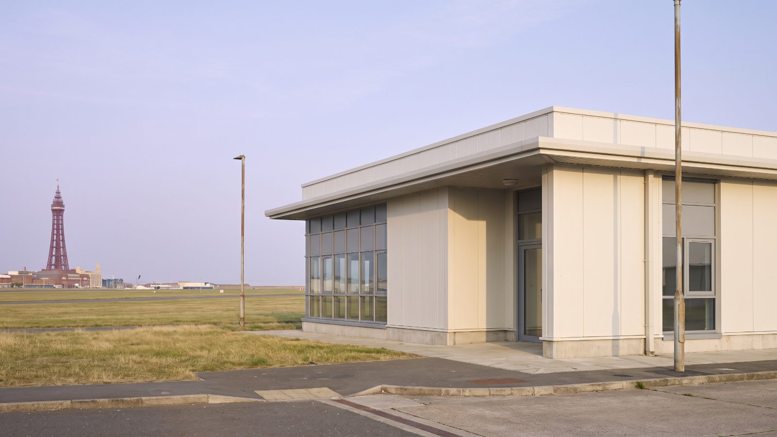 A modern commercial unit on the edge of an airfield, pale cladding, a flat grass airfield and a tall red iron lattice seaside tower on the skyline.