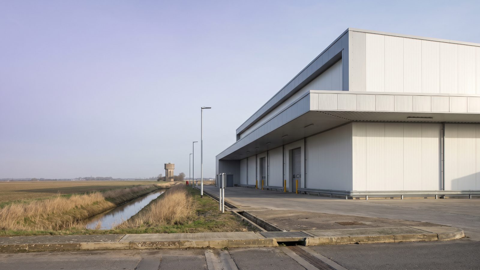 A large vegetable packhouse with white insulated cold-store panels, a vast flat landscape and wide pale sky, a long straight drainage ditch.