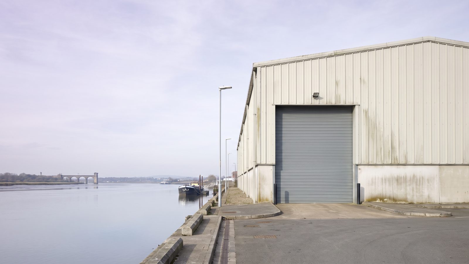 A riverside industrial unit beside a wide tidal river, pale cladding and a large door facing the water, a moored barge.